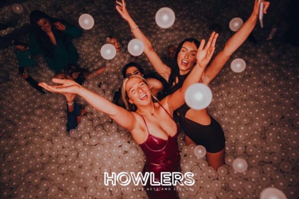 Three gorgeous girls standing in the adult ball pool in howlers with big smiles . they are throwing the balls into the air.