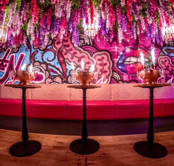 three high top tables with glasses and silver ice buckets, the bottles in the ice bucket have sparklers attached. the pick seating behind is topped with a graffiti wall with prominent pink lips. hanging down from the ceiling are are wisteria flowers.