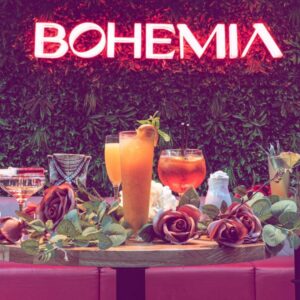 a table full of cocktails and flowers with a backdrop of a green nature wall with a neon sign for Bohemia Newcastle.