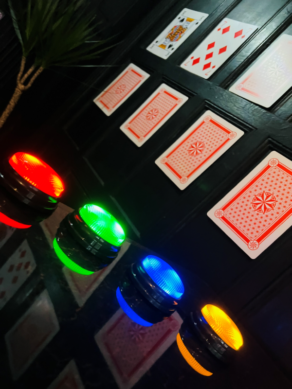 four colored buzzers in the foreground with large playing cards up on the wall in 2 rows some showing the front with a king and 7 the others turned to only show the back.