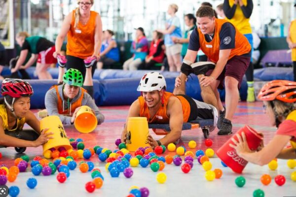 four people people with cycle helmets, lying on a flat trolly. they are holding buckets and trying to catch colored balls in the buckets while people are pushing them back and forward.