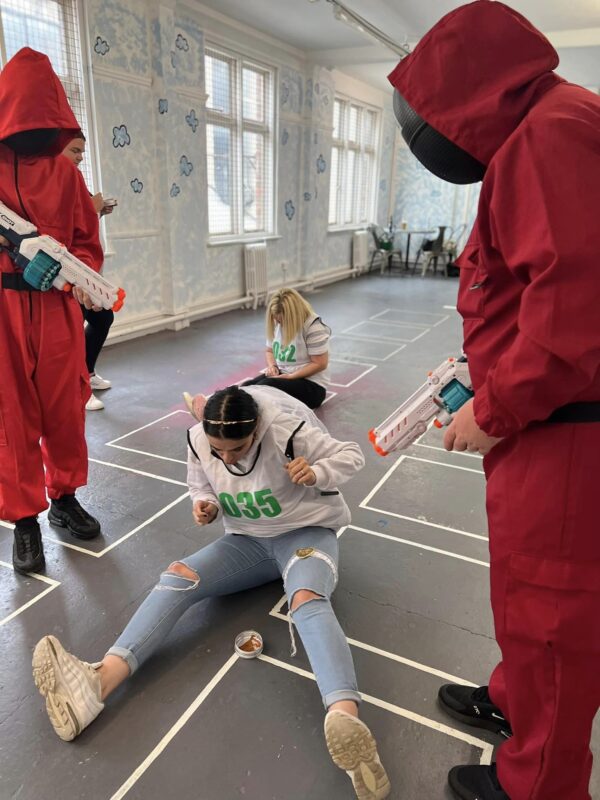 2 squid game soldiers holding guns ready to fire at a contestant. the contestant is sitting on the floor with their number bib and honeycomb tin.