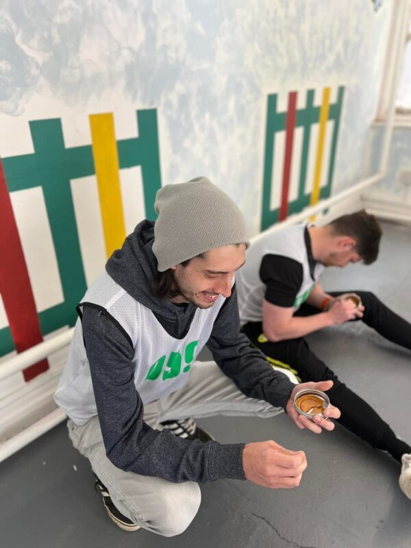 2 men sat next to a wall that has picket fences painted on it. Both are wearing squid game bibs and holding the honeycomb tins from the show.