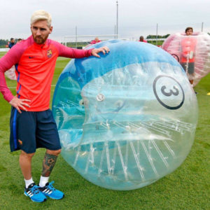 Lionel Messi Standing in his Barcelona training kit resting on a bubble football with team mates in the background standing in and leaning on Bubbles.