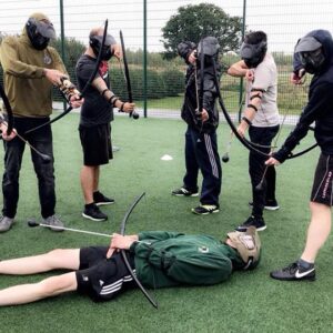 a group of men with Battlezone archery bows and arrows all loaded and pointing to a man lying on the floor.